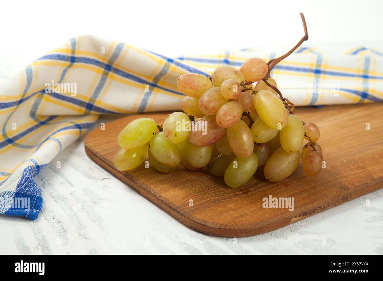 Tagliere con grande mazzo di uva rosa matura su bianco. Uva da vino rosa con asciugamano da cucina blu su fondo di legno bianco. Foto Stock