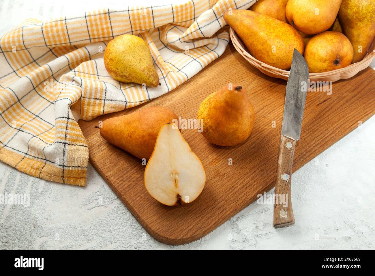 Tagliere in legno con interi e metà di succosa frutta di pera matura, cesto di vimini con pere e asciugamano da cucina giallo su fondo di legno bianco. Foto Stock