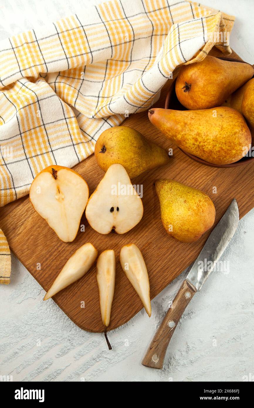 Tagliere in legno con tagliere interi, metà e fette di pere mature e succose, cesto di vimini con pere e asciugamano da cucina giallo su fondo in legno bianco Foto Stock