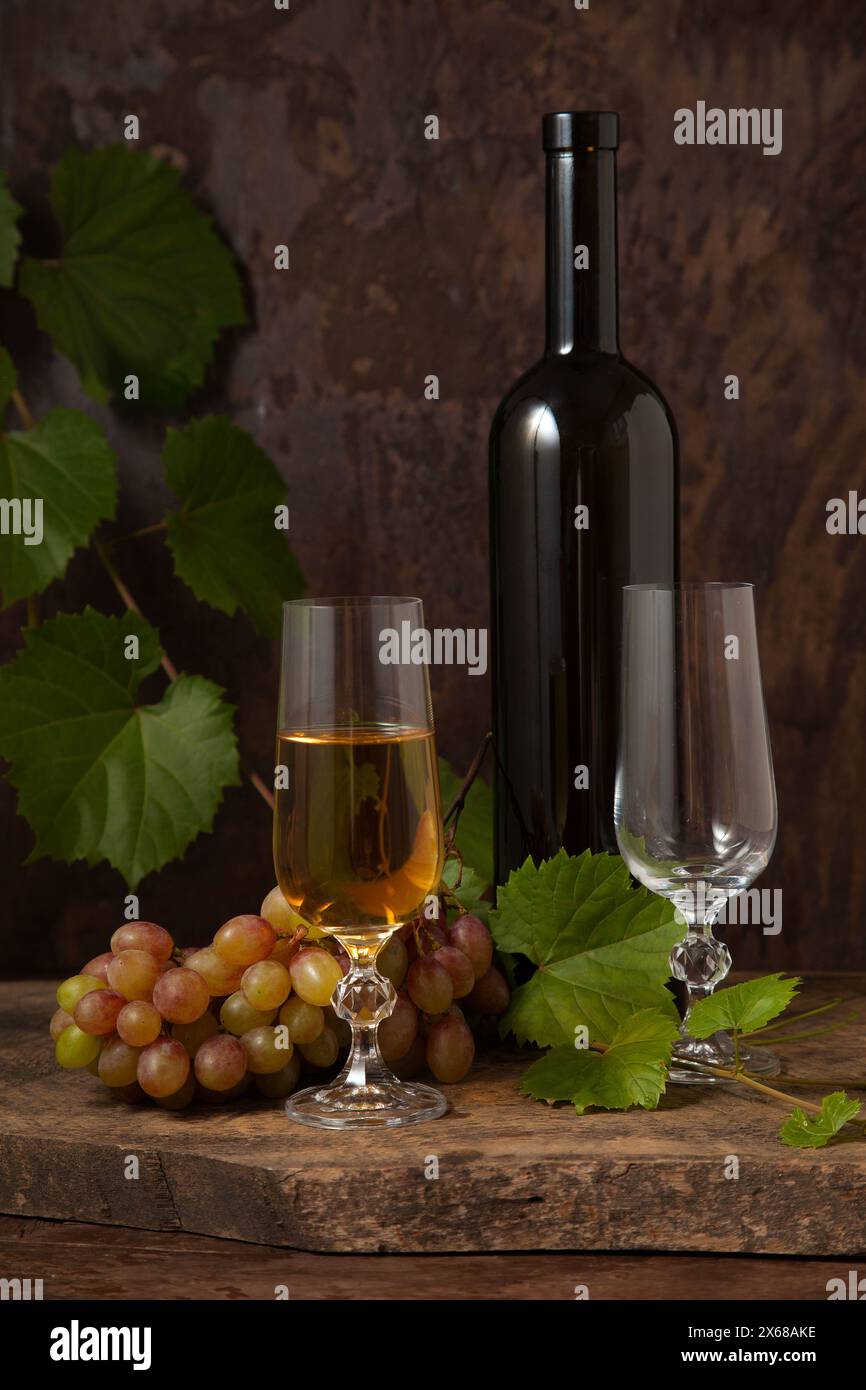 Il concetto di una deliziosa bevanda alcolica, vino e uva. Bottiglia di vino, bicchiere vuoto e bicchiere di vino bianco e mazzo di uva rosa su rustico scuro Foto Stock