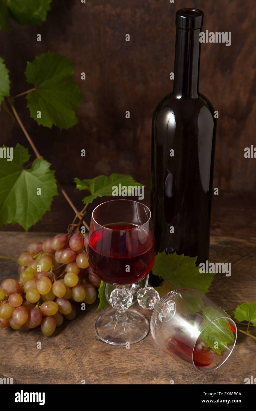 Il concetto di una deliziosa bevanda alcolica, vino e uva. Bottiglia di vino, mazzo di uva, bicchiere vuoto e bicchiere di vino rosso su legno rustico scuro Foto Stock