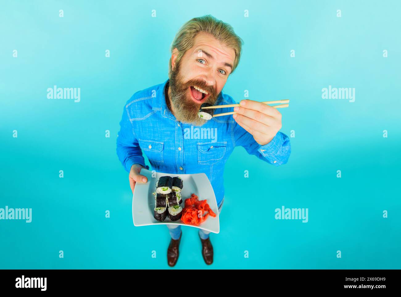 Uomo barbuto che mangia un gustoso sushi roll con bacchette di bambù. Bell'uomo in camicia in denim con piatto di sushi roll e zenzero sottaceto. Consegna di sushi Foto Stock