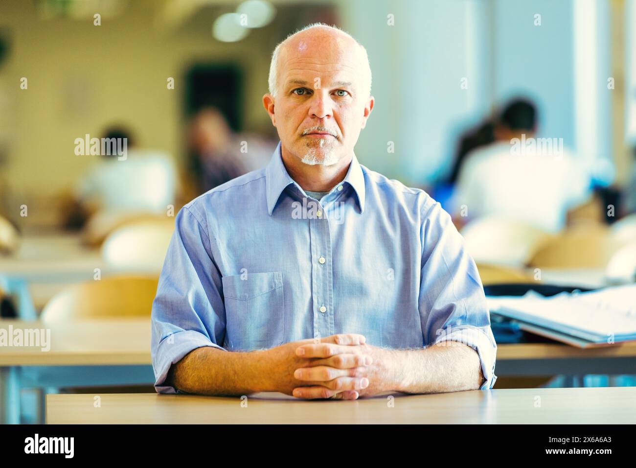 un uomo anziano con un'espressione seria siede a un tavolo in una stanza affollata e ben illuminata, con le mani schiacciate. Riflette su questioni future come il cambiamento climatico, il lavoro Foto Stock