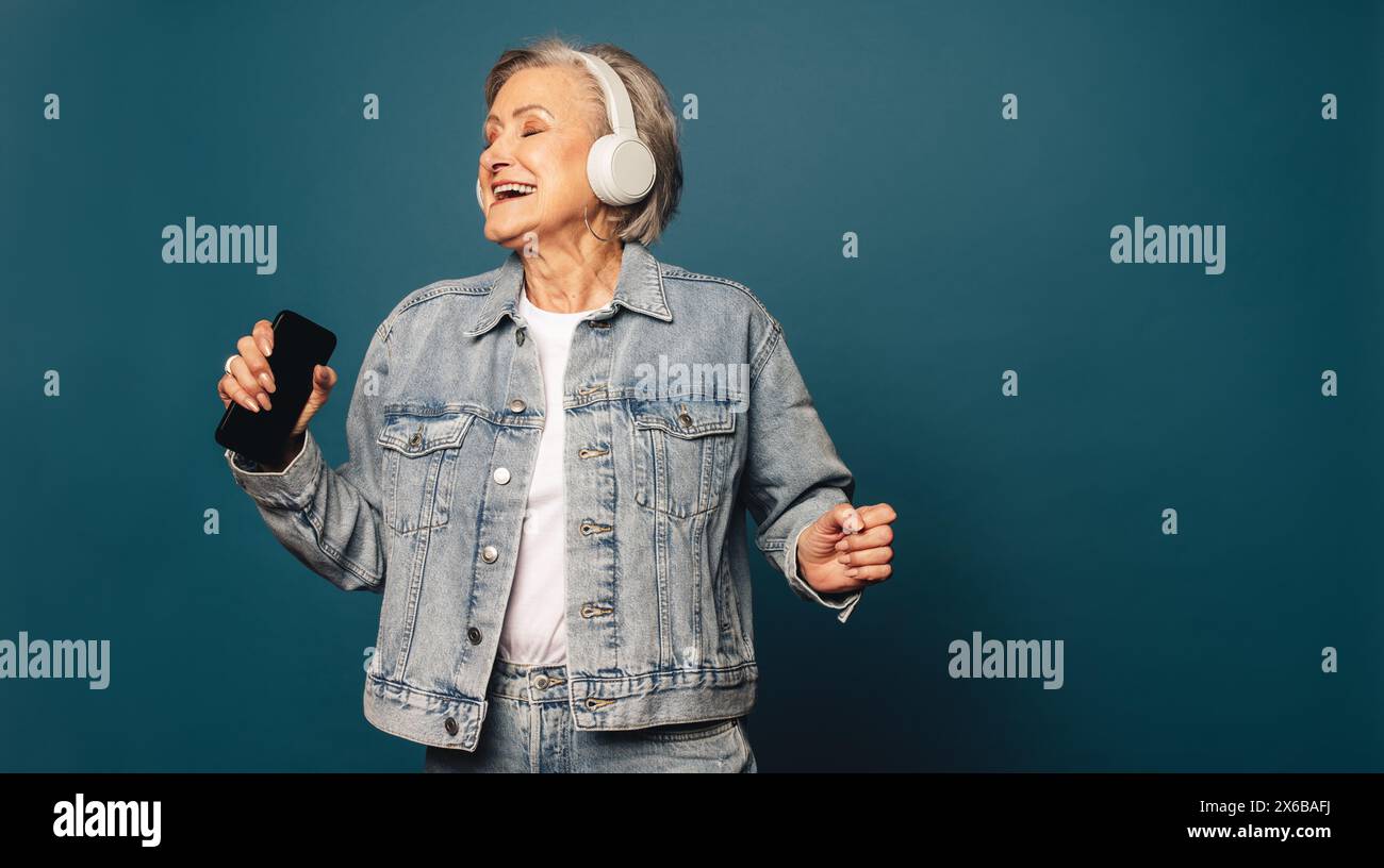Un cittadino anziano felice e alla moda in un abbigliamento casual in denim si trova in uno studio, tenendo in mano uno smartphone e indossando le cuffie. Ascolta musica, spettacolo Foto Stock