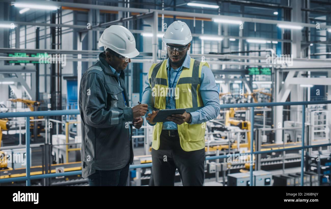 Multiethnic Manager e African American Car Factory Engineer in uniforme con tablet computer. I dipendenti del settore automobilistico 4,0 discutono del lavoro svolto nello stabilimento di assemblaggio dei veicoli. Foto Stock