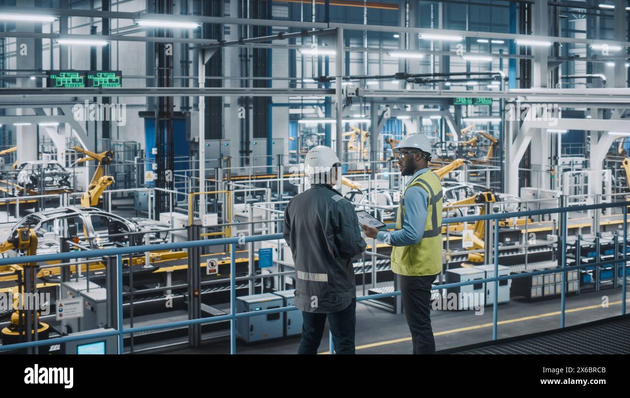 Multiethnic Manager e African American Car Factory Engineer in uniforme con tablet computer. I dipendenti del settore automobilistico 4,0 discutono del lavoro svolto nello stabilimento di assemblaggio dei veicoli. Foto Stock