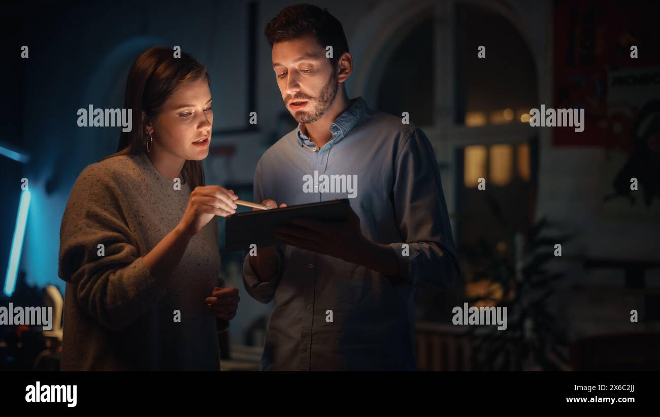 Dipendenti soddisfatti e di successo che discutono progetti correlati al lavoro su un tablet computer in Creative Agency in Loft Office. Spazio oscuro ristrutturato la sera con piante, poster e finestre arrotondate. Foto Stock