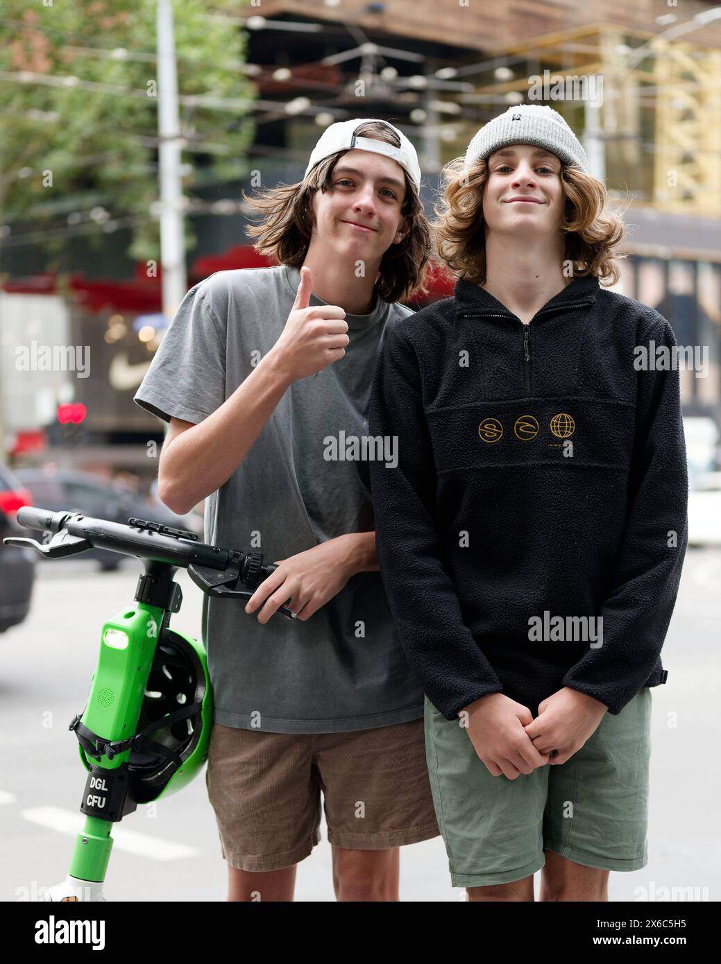 Due giovani uomini caucasici, o ragazzi, con i capelli lunghi in posa accanto a uno scooter elettrico Lime, uno che dà un gesto con i pollici in alto Foto Stock