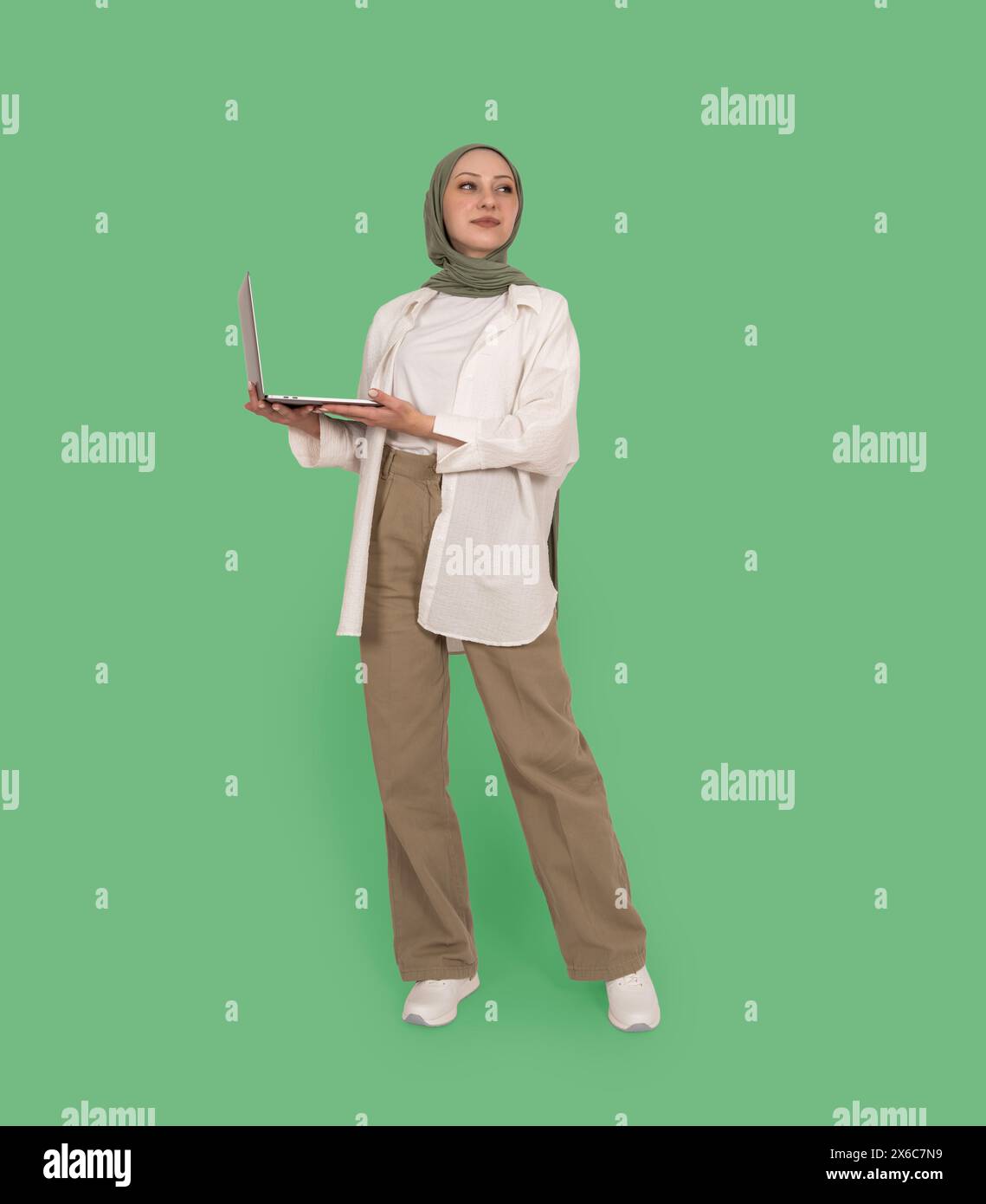 Vista del corpo a tutta lunghezza giovane donna musulmana caucasica in hijab Hold Use lavoro su laptop. Guardate da parte lo studio isolato di sfondo verde. Islam religioso. Foto Stock