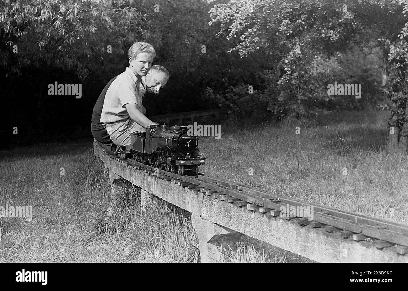 anni '1960, storico, all'esterno in un'area erbosa di un grande giardino, un giovane uomo con suo padre in sella a un treno a vapore in miniatura su una ferrovia in cemento rialzato. Foto Stock