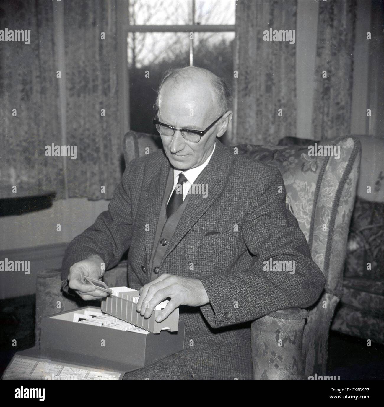anni '1960, storico, un anziano gentiluomo in costume e cravatta seduto su una poltrona che guarda le diapositive di pellicola da 35 mm contenute in un vassoio Le diapositive fotografiche risalgono al 1935, quando Kodak introdusse la pellicola slide Kodachome, che iniziò rapidamente il formato cinematografico più popolare di tutti i tempi. le diapositive da 35 mm sono anche denominate lucidi fotografici perché dispongono di una finestra che consente al visualizzatore di vedere la pellicola fisica. Foto Stock