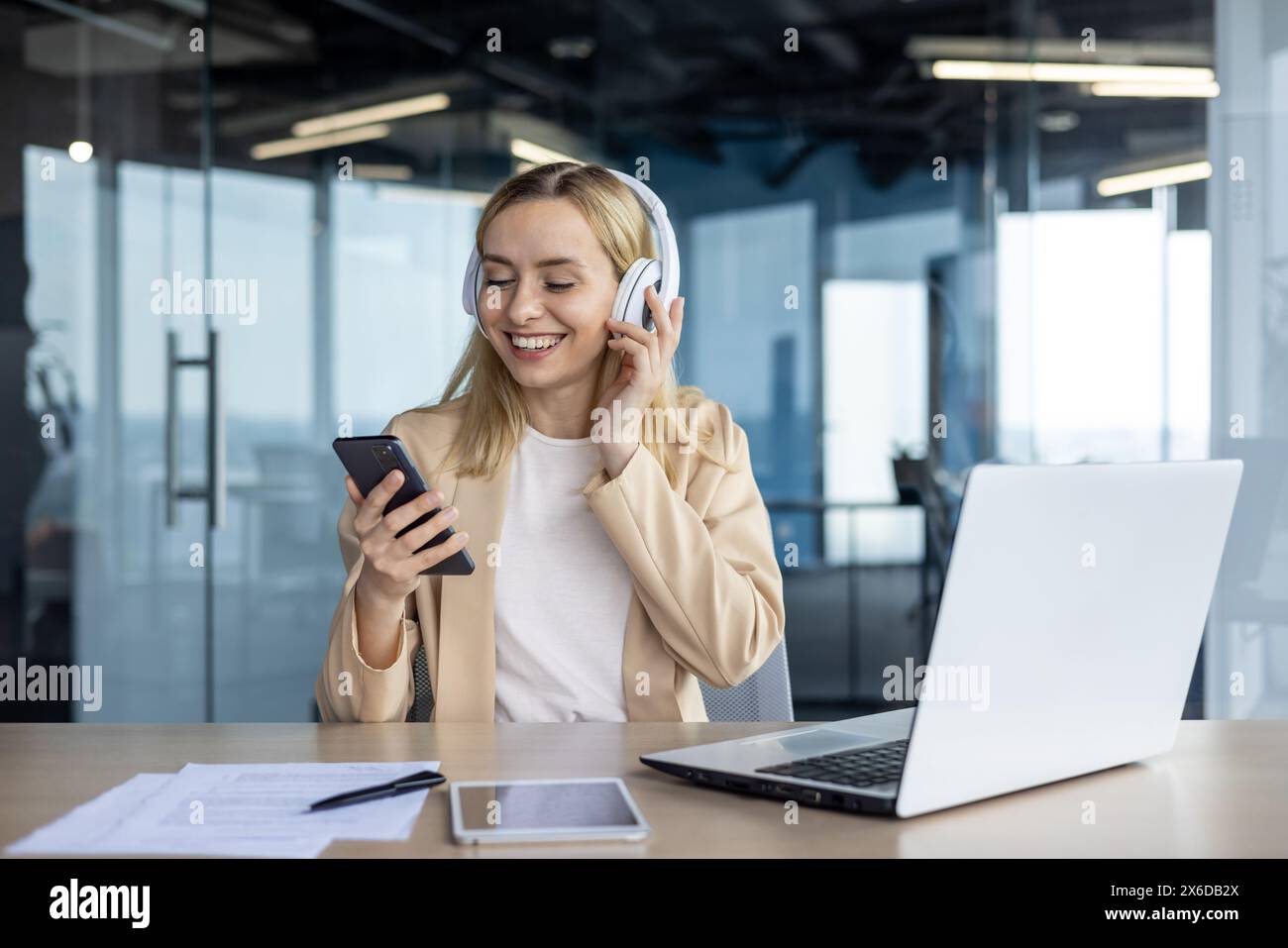 Una giovane donna d'affari felice ascolta la musica con le cuffie mentre utilizza lo smartphone e il computer portatile in un ambiente luminoso. Una piccola pausa durante il lavoro. Foto Stock