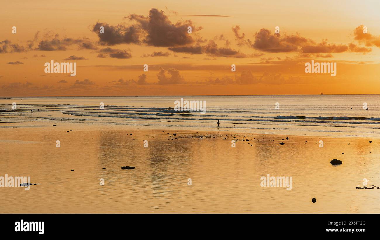 Sfumature dorate dipingono il cielo sulla spiaggia di Bingin, Bali, mentre il sole scende sotto l'orizzonte, proiettando un tranquillo bagliore sulle onde e sulle silhouette della sera Foto Stock
