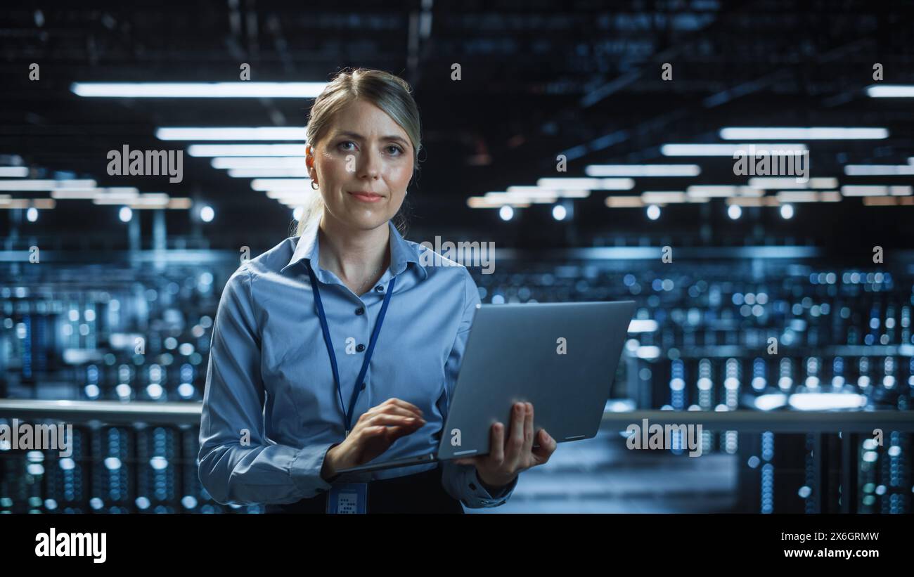 Ritratto di uno specialista IT sorridente che utilizza un computer laptop nel data center, guarda la fotocamera. Ingegnere e-business di successo che lavora in una struttura di cloud computing per aziende agricole di grandi server. Foto Stock