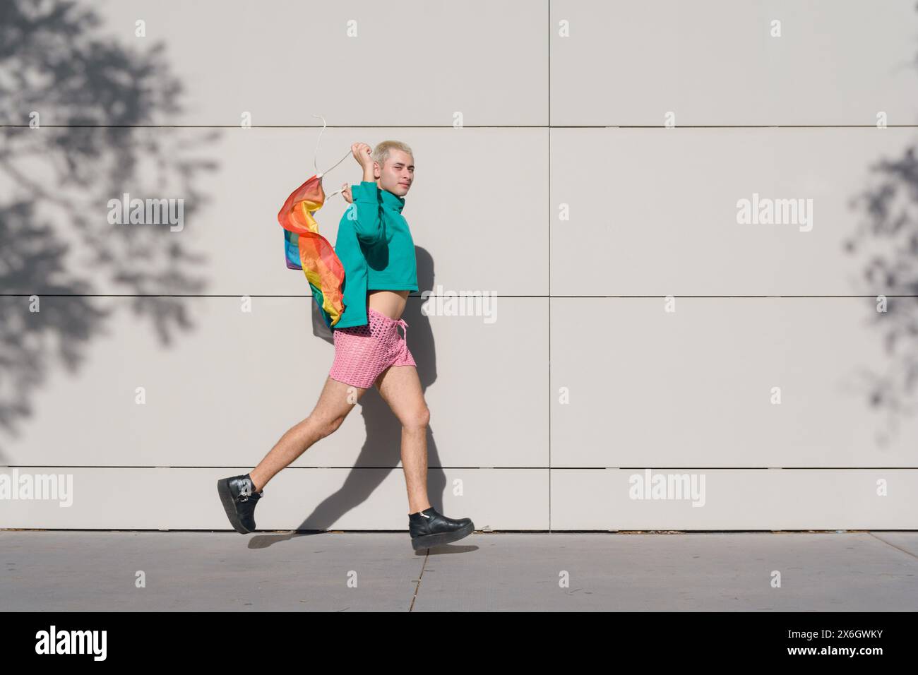 Il giovane latino con i capelli biondi corti, con pantaloncini rosa, sneakers nere e maglione verde, è all'aperto con la bandiera dell'orgoglio e si gode il tramonto Foto Stock