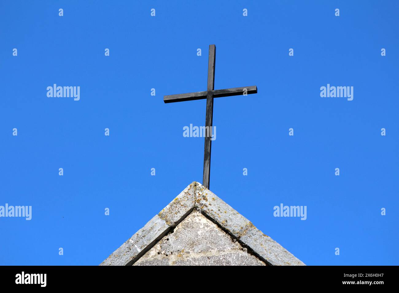 Vecchia croce di metallo nero, logora e fatiscente, sulla parte superiore della tradizionale chiesa cattolica in pietra parzialmente ricoperta di muschio giallo sul retro del cielo azzurro Foto Stock