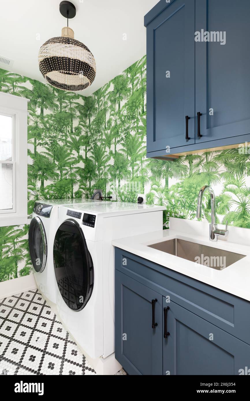 Un'accogliente lavanderia con armadietti blu, ripiano in marmo bianco, cestello luminoso appeso sopra la lavatrice e l'asciugatrice e carta da parati verde e bianco. Foto Stock