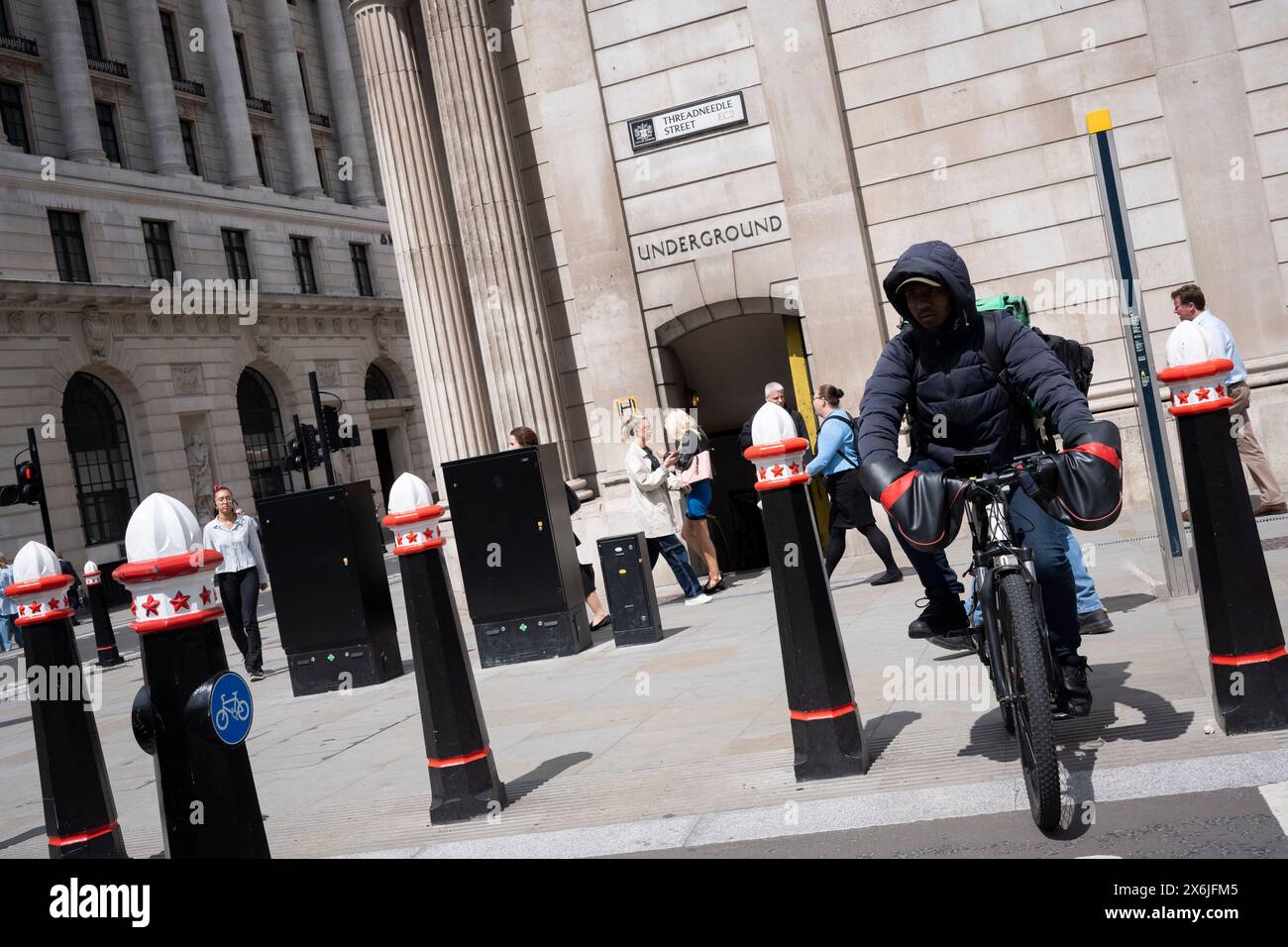 A causa della pista ciclabile chiusa durante i cambiamenti di disposizione delle strade, un ciclista di Uber Eats passa all'ora di pranzo i lavoratori della City camminano oltre la Bank of England all'incrocio con la City of London's Bank, nel cuore del quartiere finanziario della capitale, il 15 maggio 2024, a Londra, Inghilterra. Foto Stock