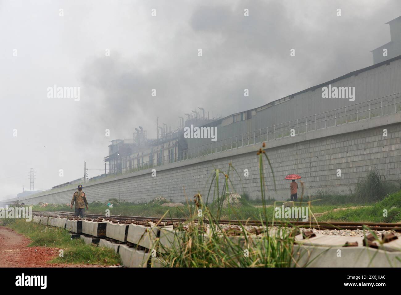 Dacca, Bangladesh. 15 maggio 2024. La gente passa un'area industriale mentre il fumo sale dai mulini rilaminati a Shyampur, a Dacca, Bangladesh, 15 maggio 2024. Secondo uno studio della Banca mondiale, il Bangladesh deve affrontare un elevato livello di inquinamento atmosferico, che costa circa il 9% del PIL all'anno. La maggior parte dei laminatoi in acciaio all'interno e intorno alla capitale sono stati eseguiti senza il necessario sistema di controllo dell'inquinamento atmosferico che poneva un rischio di inquinamento atmosferico e di pericolo per la salute. Foto di Suvra Kanti Das/ABACAPRESS. COM credito: Abaca Press/Alamy Live News Foto Stock