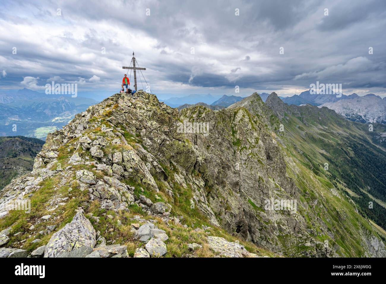 Alpinista sulla cima rocciosa a punta del Raudenspitze o Monte Fleons con croce sommitale, panorama montano sul crinale principale del Carnico, Carnic Foto Stock