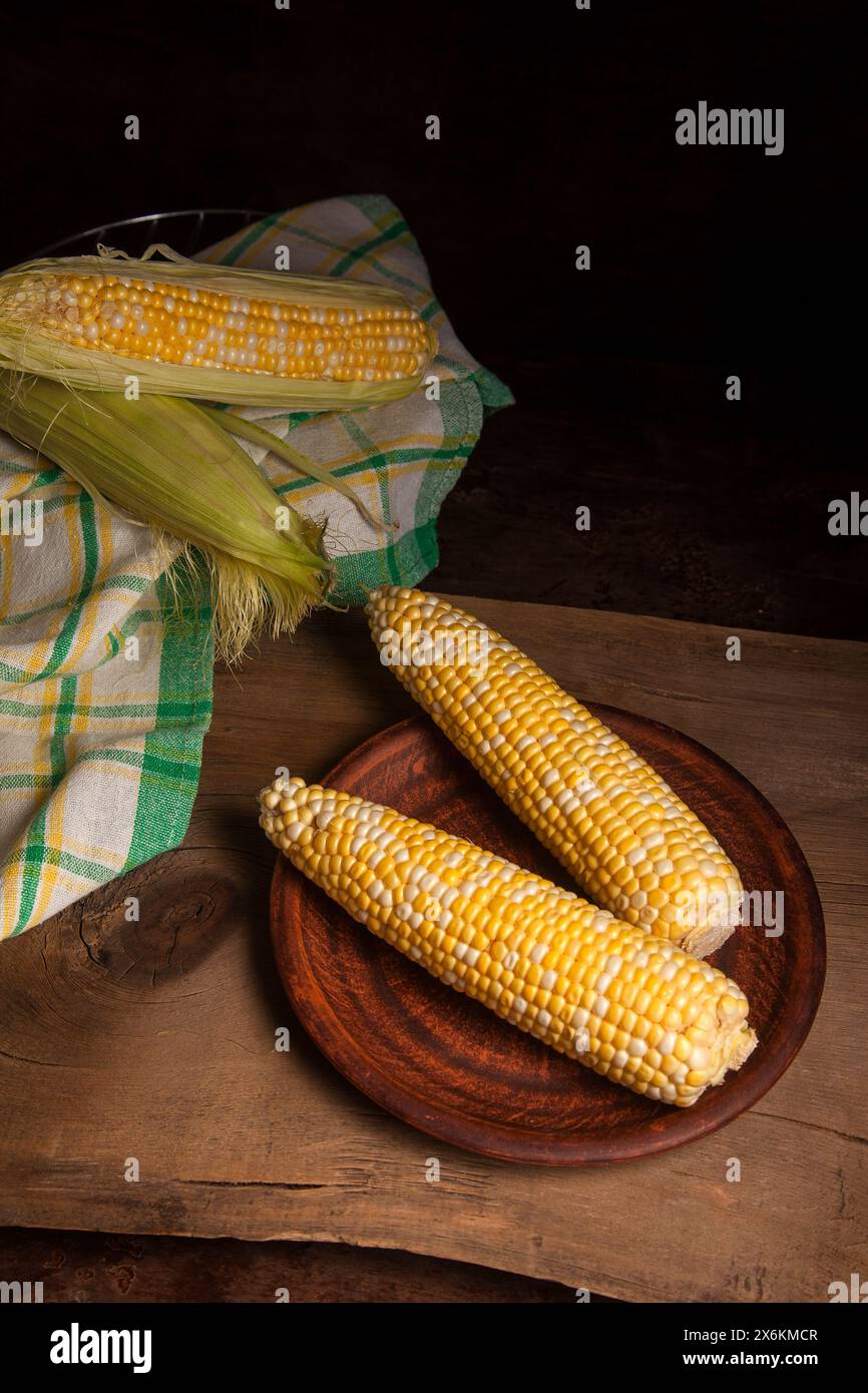 Piatto di argilla con due spighe di mais dolce maturo e asciugamano verde su fondo in legno vintage. Pannocchie con grani bianchi e gialli. Spighe fresche di mais con g Foto Stock