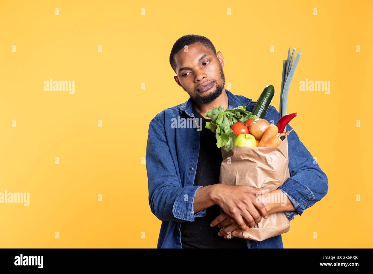 Un ragazzo pensivo e dispiaciuto tiene in mano un sacchetto di carta pieno di cibo biologico, acquistando frutta e verdura ecologiche. Delusa persona afroamericana triste acquista prodotti biologici. Foto Stock