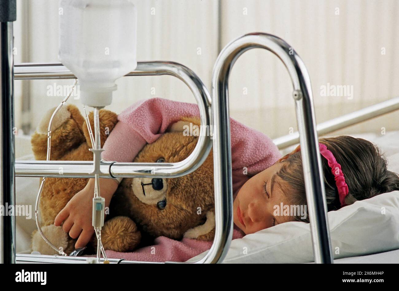Una giovane ragazza dorme tranquillamente in un letto d'ospedale, confortata da un orsacchiotto al suo fianco. La ragazza sembra riposare pacificamente Foto Stock