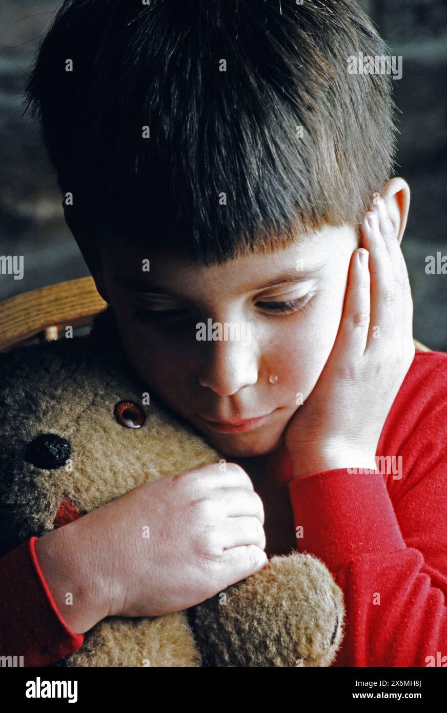 Un ragazzo lacrime e infelice che indossa una camicia rossa viene mostrato mentre abbraccia un orsacchiotto mentre è seduto tranquillamente in casa. Sembra che stia cercando conforto e lo è Foto Stock