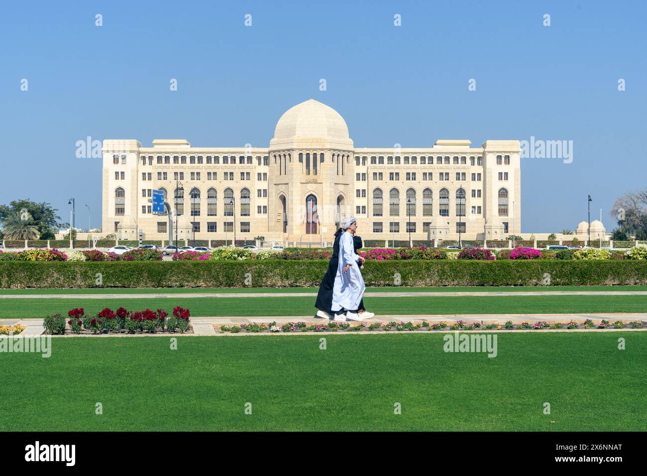 Mascate, Oman - 2 gennaio 2024: Una figura solitaria fa una tranquilla passeggiata nel verde lussureggiante, con la maestosa architettura di un grande edificio Foto Stock