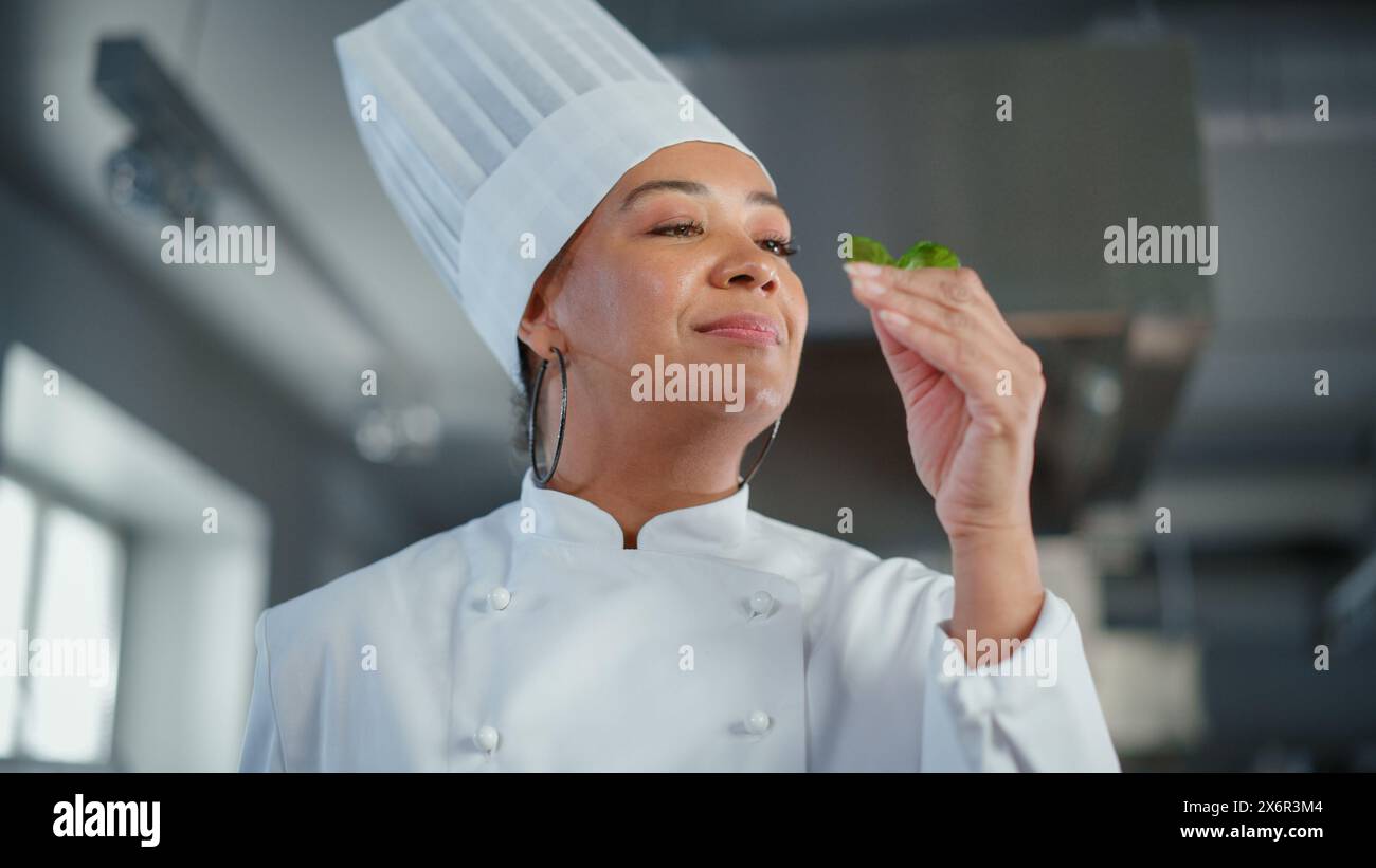 Black Female Chef prende le erbe fresche, le profuma con un sorriso, ingrediente segreto che rende speciale la ricetta della nonna. Cucina tradizionale del ristorante con piatti autentici, cibo salutare e nutriente Foto Stock