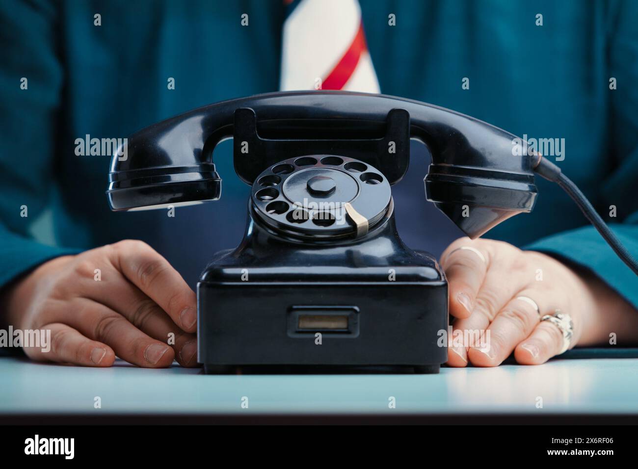Appoggiate le mani accanto a un vecchio telefono rotativo nero su una scrivania, la persona che indossa una giacca color ottanio e una cravatta a righe, suggerendo di essere pronta a comunicare Foto Stock