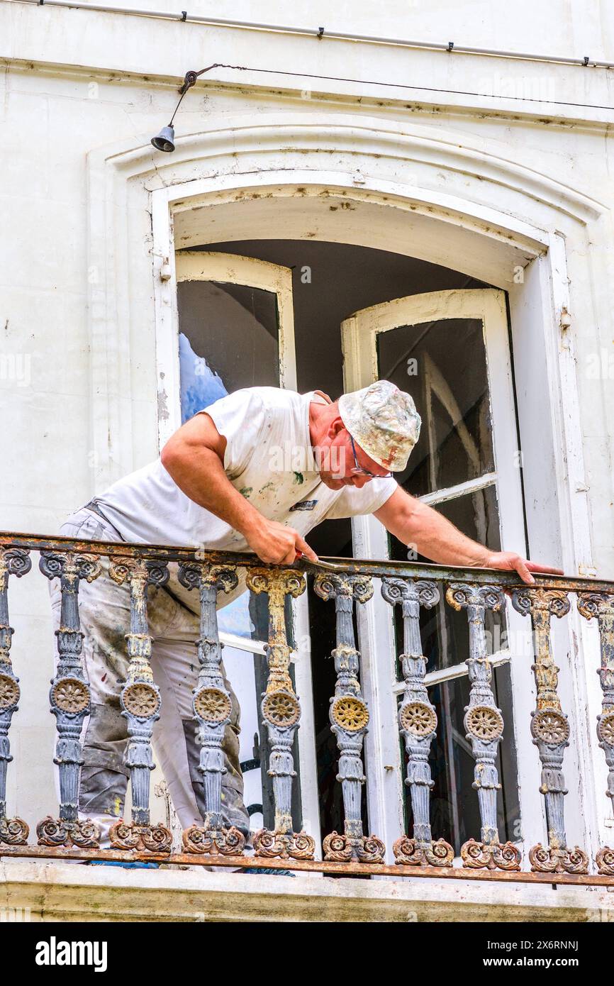 Lavoratore che utilizza una spazzola metallica per raschiare la vernice e la ruggine da ringhiere in ferro ghiacciato per balconi - Loches, Indre-et-Loire (37), Francia. Foto Stock