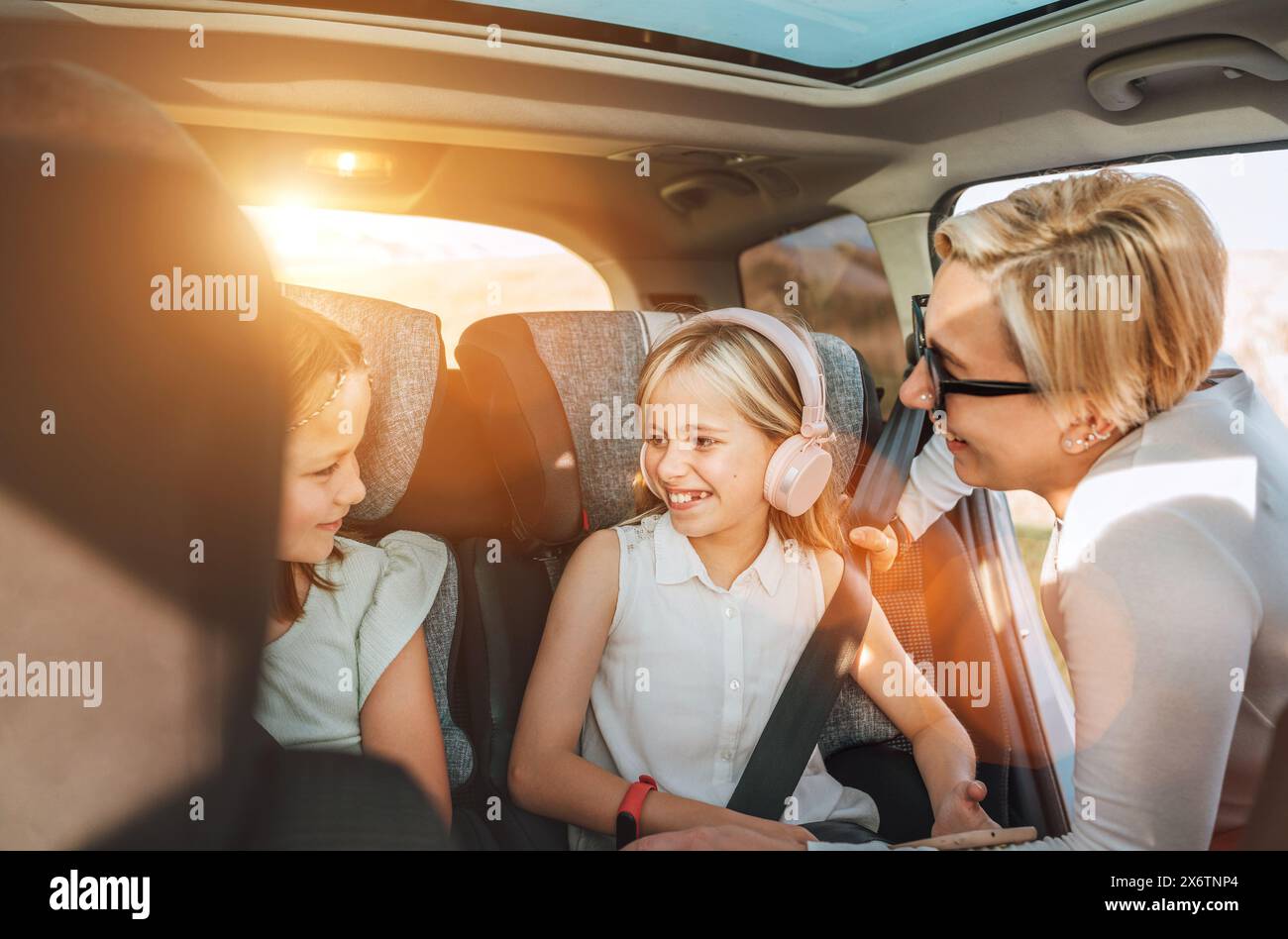 All'interno dell'auto foto di una madre che si stringe con la cintura di sicurezza della bambina seduta sul seggiolino per bambini. Ragazza che ascolta musica con le cuffie Foto Stock
