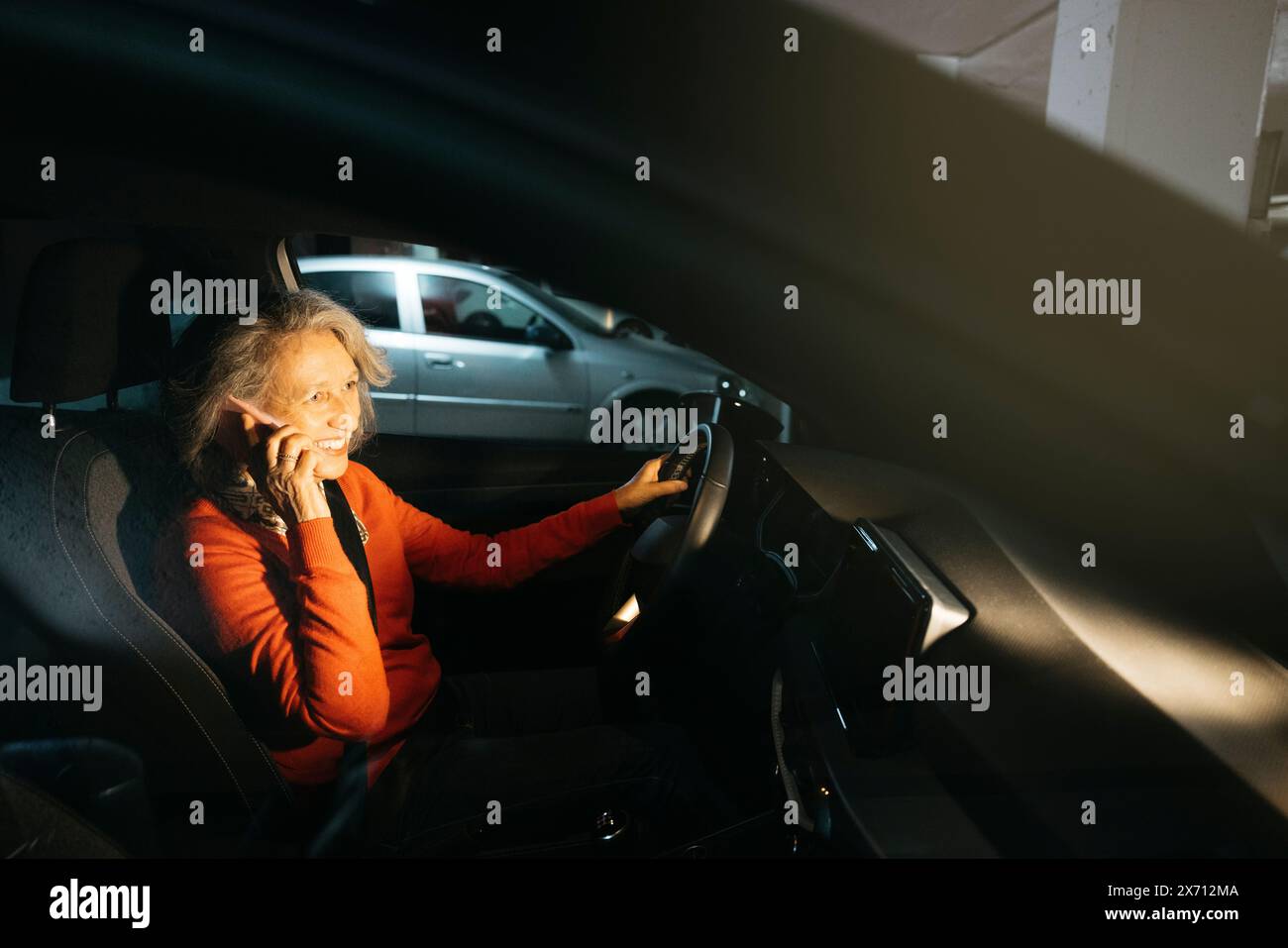 Donna anziana seduta in un'auto parcheggiata in un garage sotterraneo che sorride mentre parla al telefono. Foto Stock