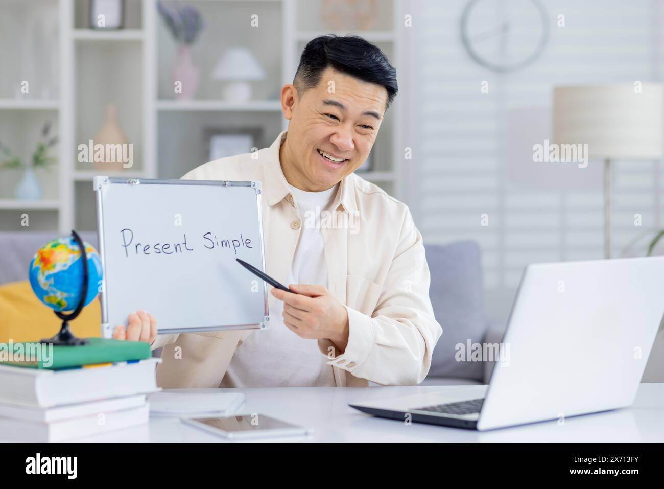Un insegnante di lingue online sorridente che spiega di presentare un semplice tempo durante una lezione virtuale. Uomo che tiene una lavagna bianca e punta con una penna durante l'insegnamento tramite laptop. Foto Stock