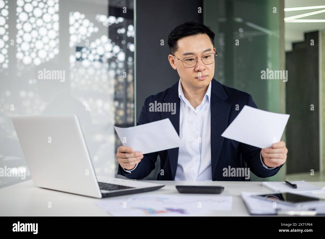 Uomo maturo asiatico alla sua scrivania in un ufficio moderno, che esamina attentamente i documenti. Ambiente professionale con laptop e documenti. Foto Stock