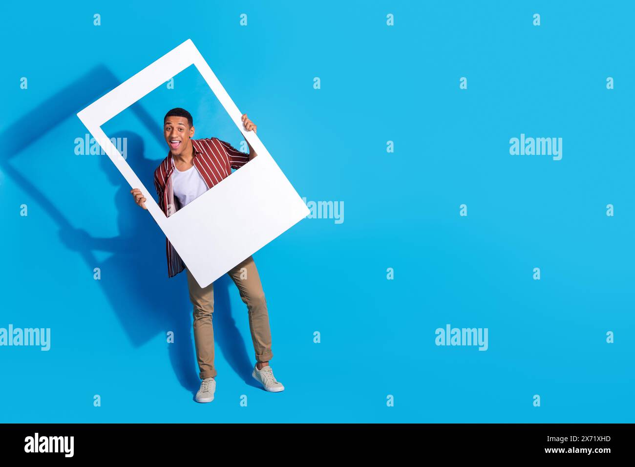 Foto a grandezza naturale di pantaloni camicia a righe dall'uomo allegro e funky, con cornice per foto isolata su sfondo blu Foto Stock