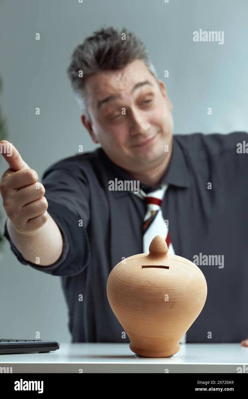 Un uomo caucasico con camicia grigia scura e cravatta a righe, con capelli grigi spigolosi, punta il pollice verso un salvadanaio in ceramica e indossa un oggetto appagante Foto Stock