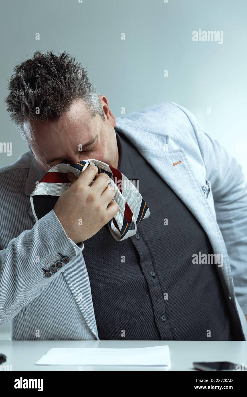 Uomo dai capelli grigi chiari con un blazer grigio chiaro, camicia grigio scuro e cravatta a righe, usa la cravatta per soffiarsi il naso, sguardo disperato, seduto a una scrivania, semplice Foto Stock