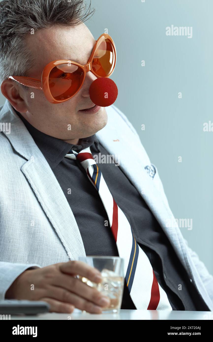 Indossando una tuta leggera e una cravatta a righe, l'uomo indossa occhiali arancioni sovradimensionati e un naso rosso clown tenendo in mano un bicchiere di whisky. Il suo stupido comportamento si aggrava Foto Stock