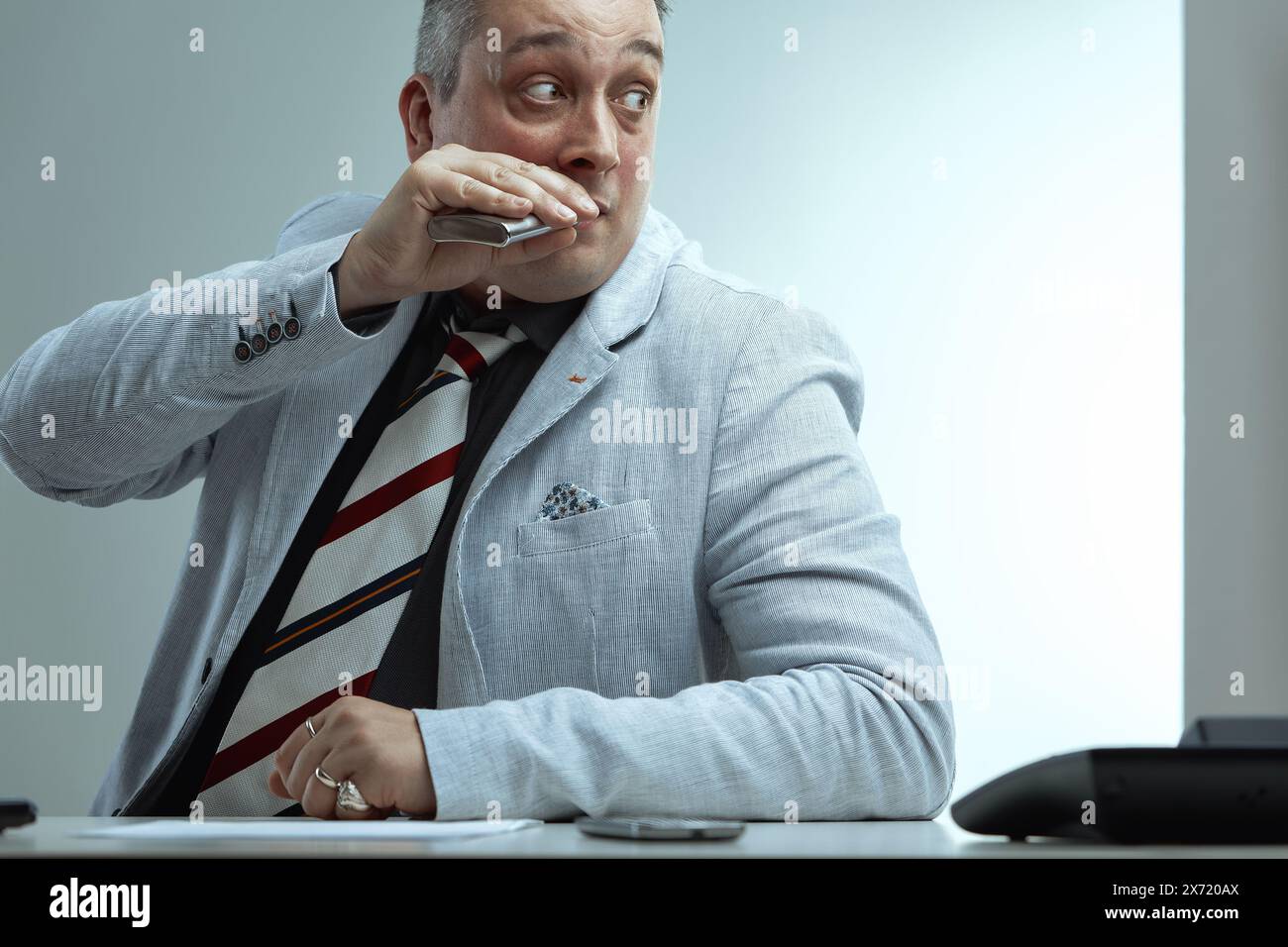 Uomo con capelli grigi spioventi, giacca da tuta grigio chiaro, camicia grigio scuro e cravatta a righe, sorseggia nervosamente da una fiasca mentre guarda lateralmente, apparendo afr Foto Stock