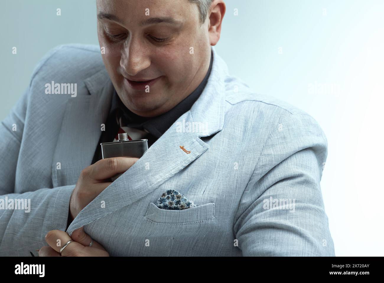 Uomo caucasico con blazer grigio chiaro, camicia grigio scuro e cravatta a righe, con capelli grigi piccanti, sorseggia nervosamente da una fiasca mentre guarda lateralmente, feari Foto Stock