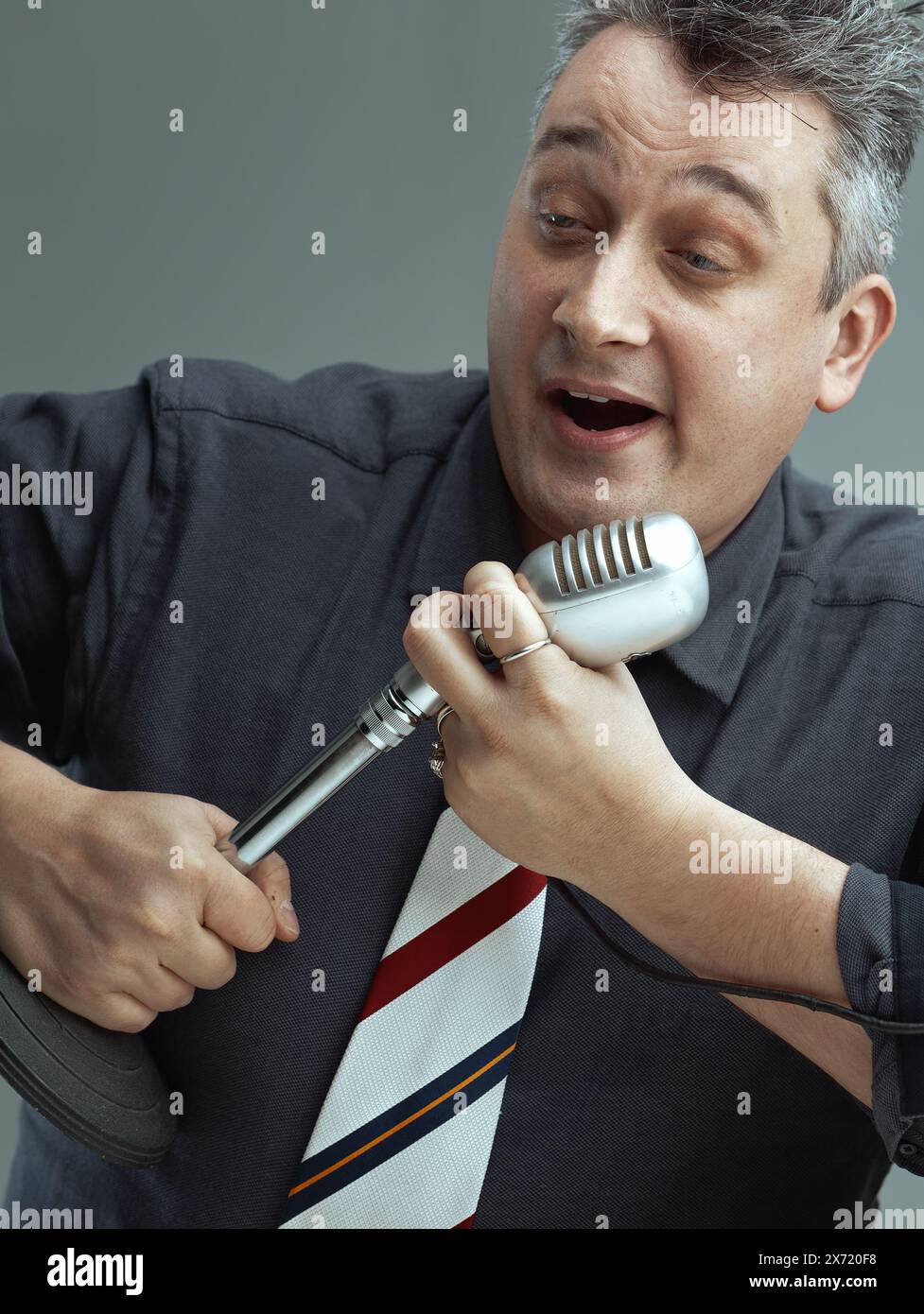 l'uomo, vestito con una camicia scura e cravatta a righe, canta energicamente in un microfono vintage. Il suo comportamento espressivo e la sua postura entusiasta mostrano il suo Foto Stock