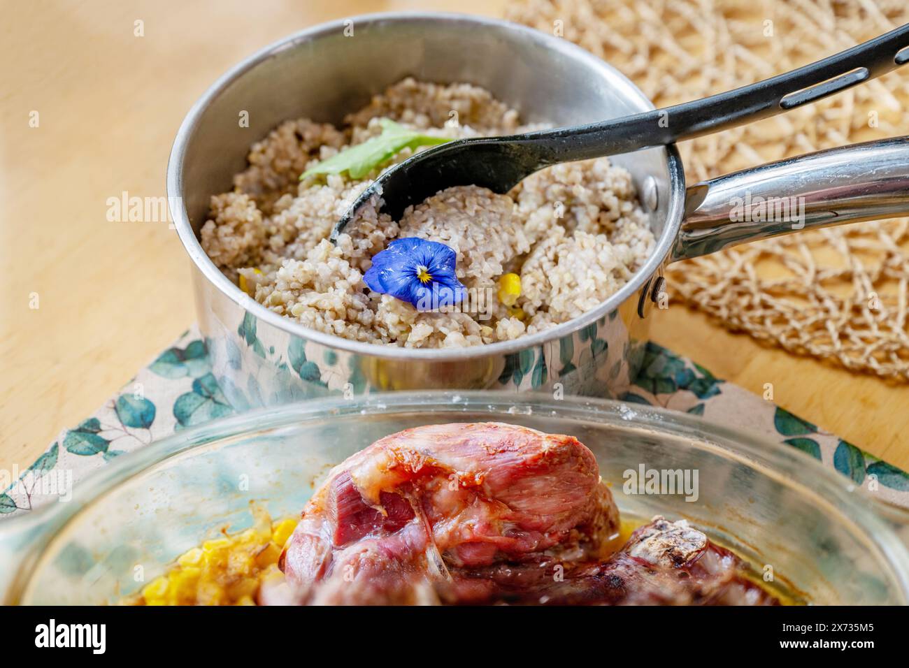 Grano saraceno bollito con fiore commestibile in pentola con cucchiaio, parte di carne di vitello cotta in pentola di vetro sul tavolo, primo piano. Foto Stock