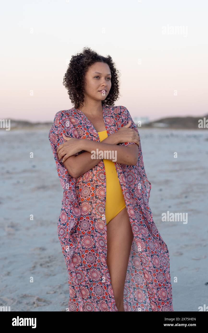 In spiaggia, giovane donna birazziale in piedi da sola, che attraversa le braccia Foto Stock