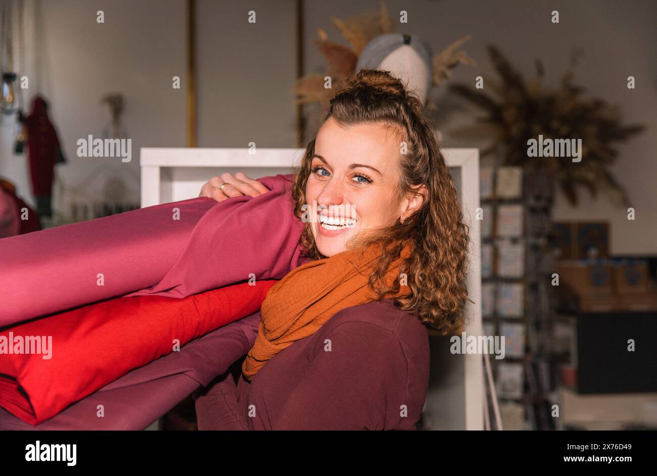 Donna amichevole con capelli ricci in un ambiente creativo da officina, che maneggia una pila di rotoli di tessuto vivaci Foto Stock