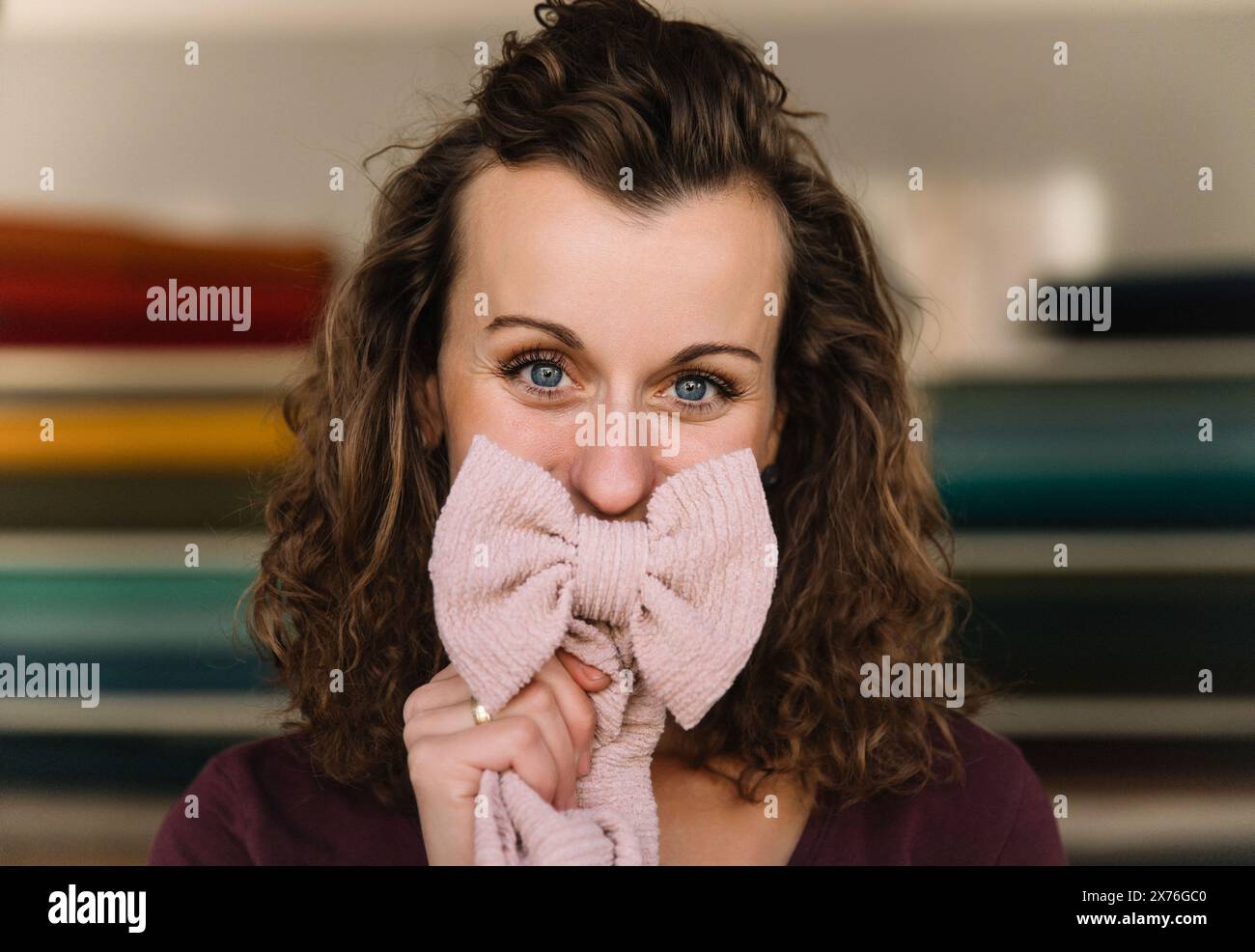 Vista ravvicinata che cattura gli occhi vivaci di una stilista che si copre giocosamente il viso con un fiocco in maglia rosa, suggerendo la sua personalità creativa Foto Stock