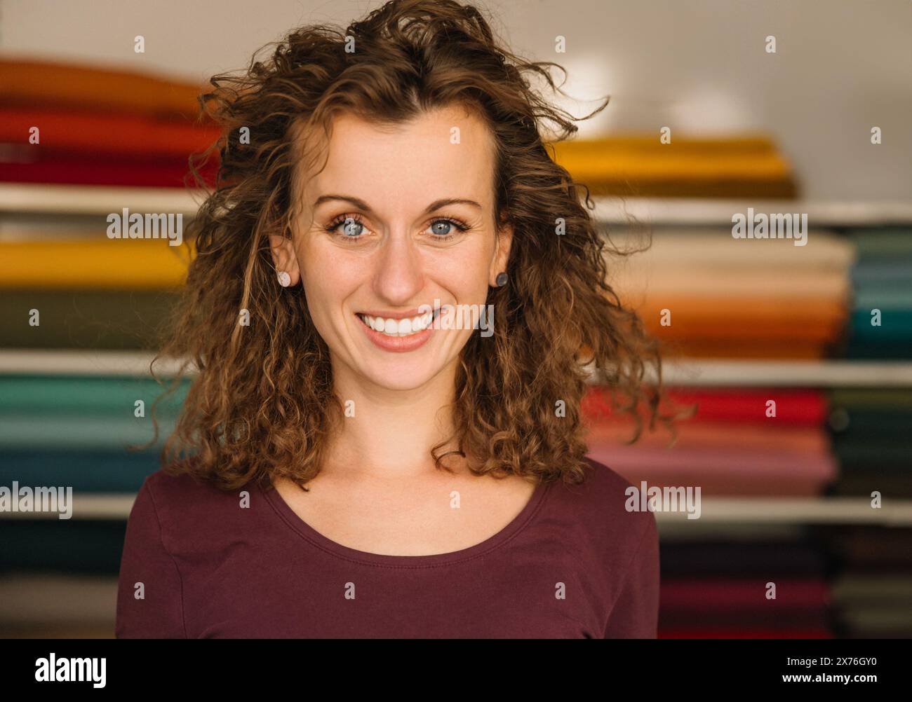 Una vivace donna dai capelli ricci sorride calorosamente nel suo colorato laboratorio tessile, mostrando la sua bellezza naturale e l'ambiente creativo Foto Stock