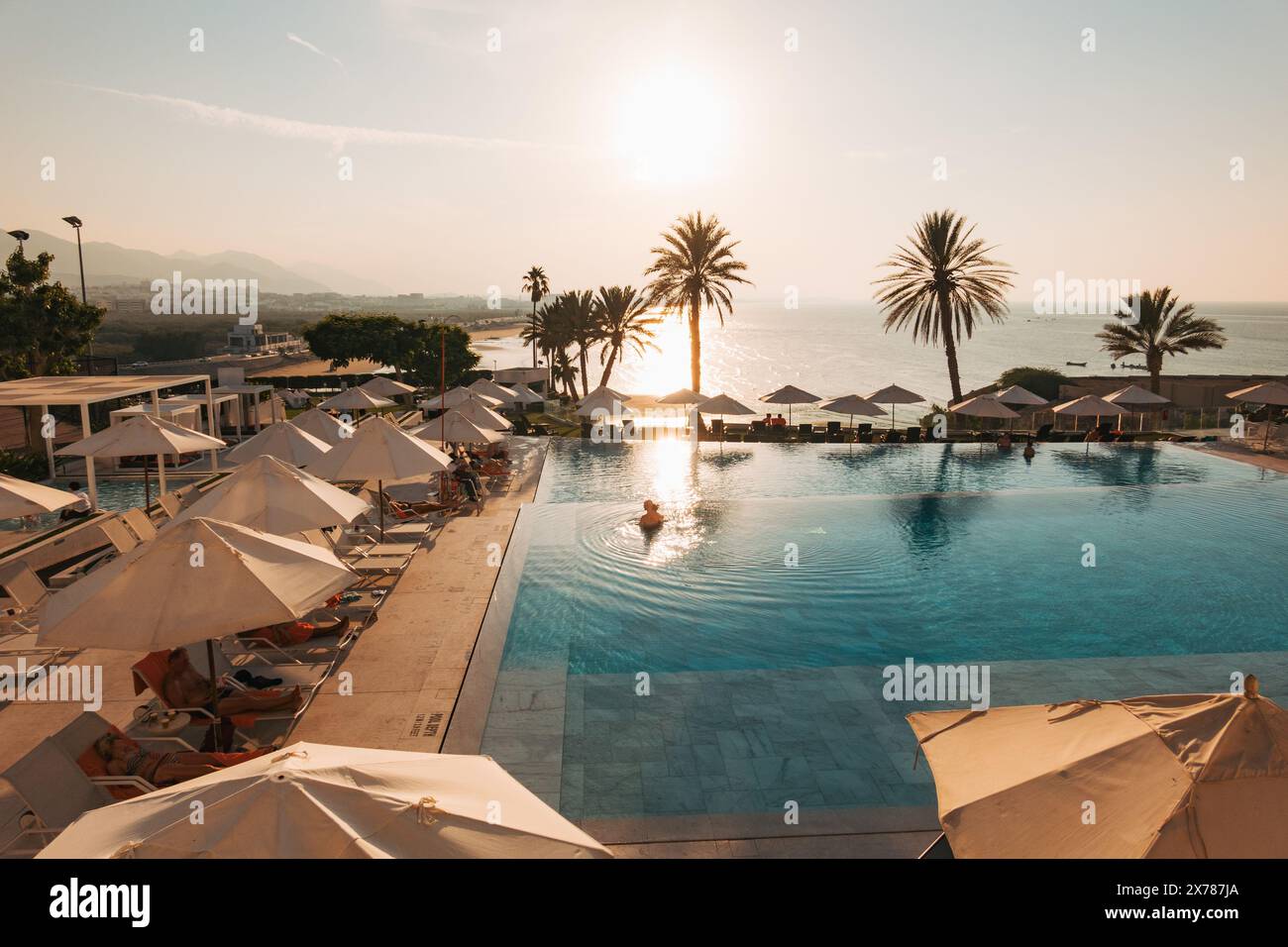 Vista del tramonto sulla piscina dell'IHG Crowne Plaza Muscat, con palme, lettini e la costa del Golfo dell'Oman sullo sfondo. Foto Stock