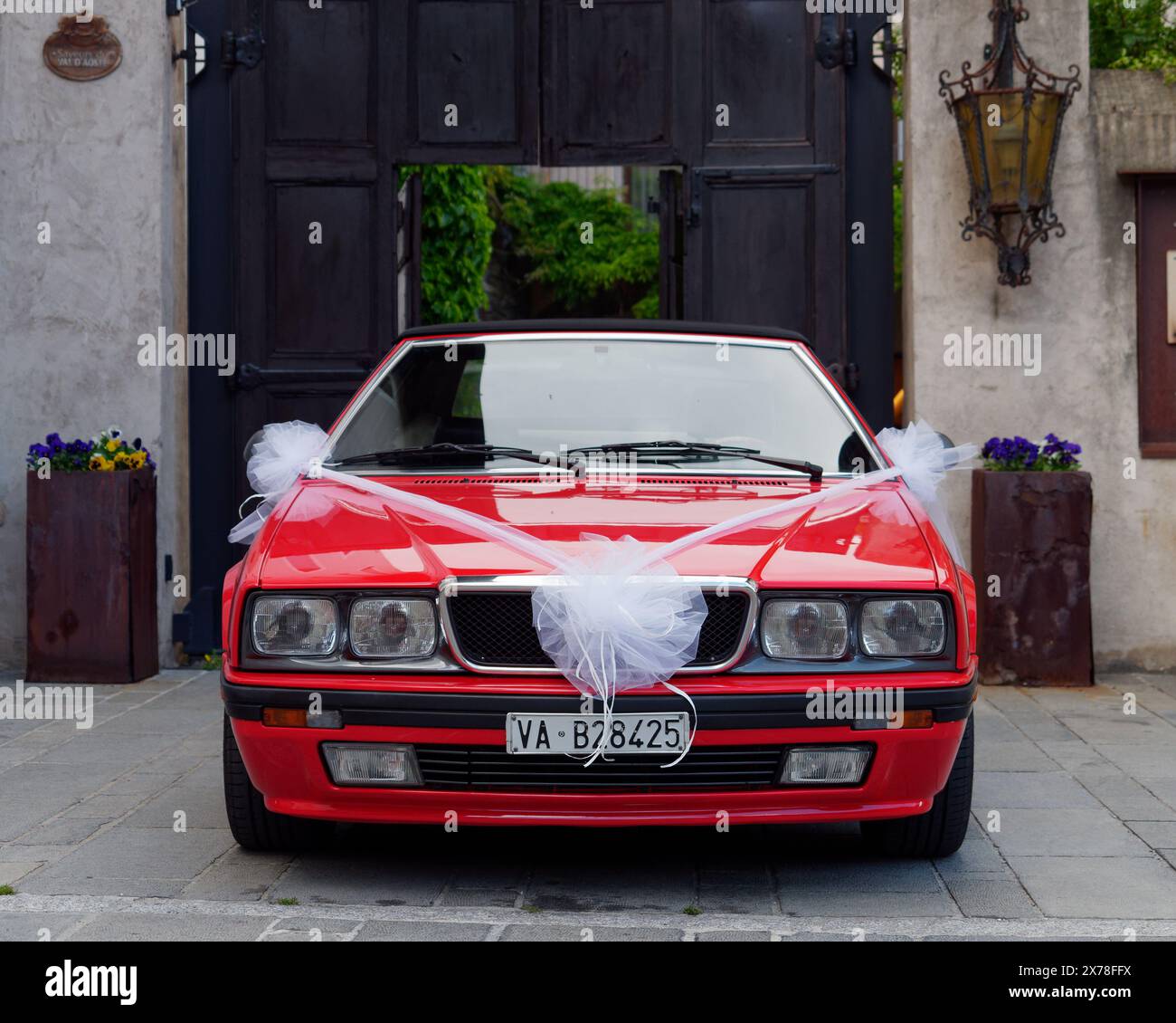 Auto da sposa rossa convertibile con nastro bianco nella città di Nus di fronte a un'elegante porta di legno. Valle d'Aosta, 18 maggio 2024. Foto Stock